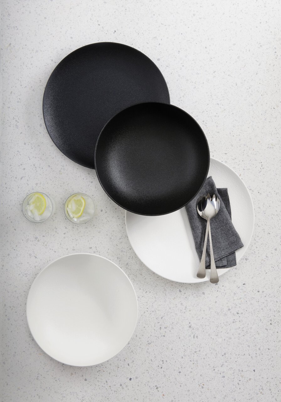 Vogelperspektive auf ein 8-teiliges Dinner-Set in Schwarz und Weiß, bestehend aus Tellern und Schalen, mit Besteck und Servietten auf einem hellen Untergrund.
