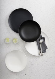 Vogelperspektive auf ein 8-teiliges Dinner-Set in Schwarz und Weiß, bestehend aus Tellern und Schalen, mit Besteck und Servietten auf einem hellen Untergrund.