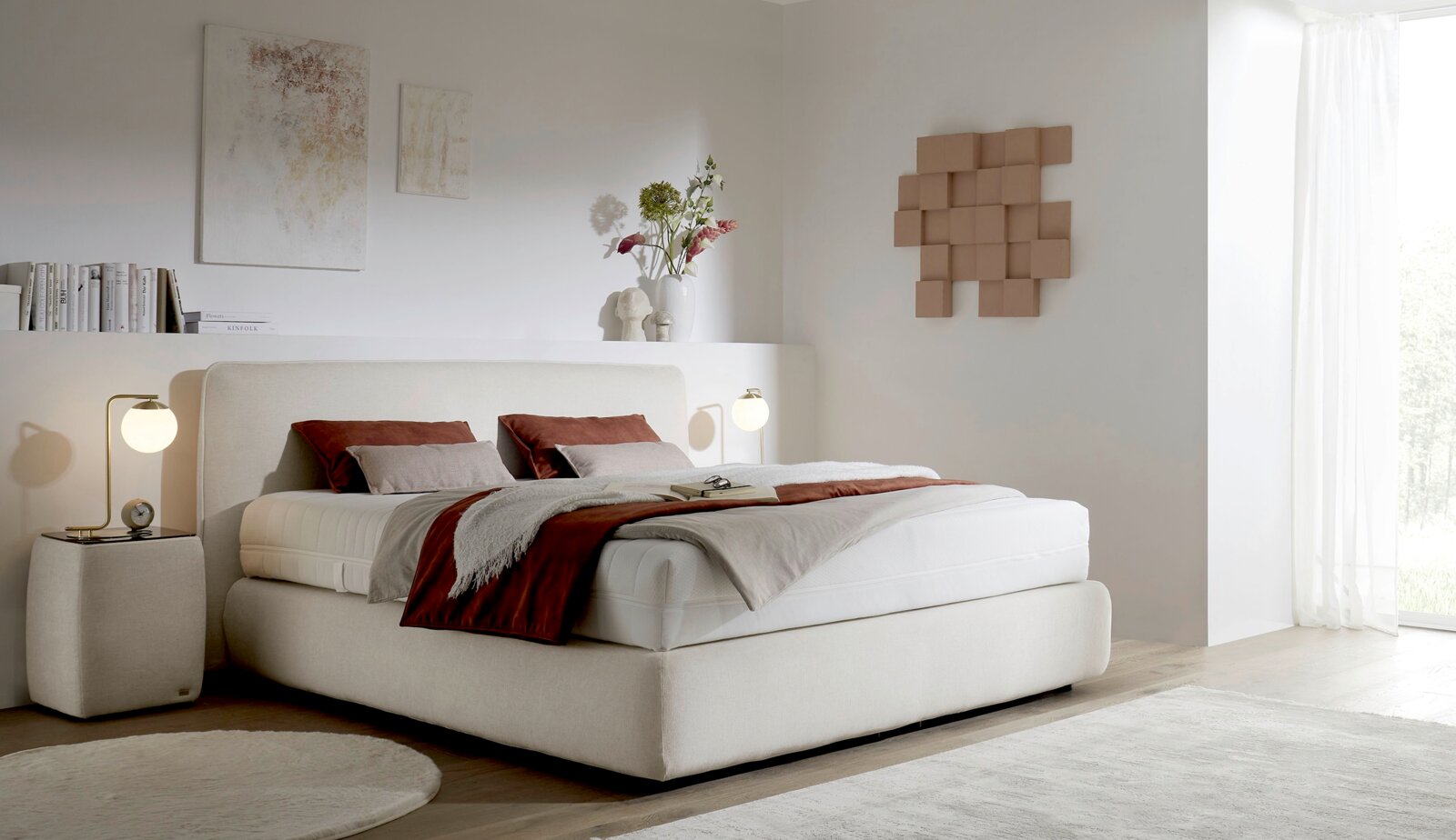 Helles, modern gepolstertes Boxspringbett in Creme mit weißer Matratze und rostfarbenen Kissen/Decke im minimalistischen Schlafzimmer, Ansicht schräg von vorne.