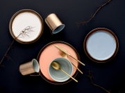 Draufsicht auf ein modernes Keramik-Geschirrset mit Tellern in Weiß, Blau und Rosé, Schale und Tassen sowie goldenem Besteck auf dunklem Hintergrund mit dekorativen Zweigen