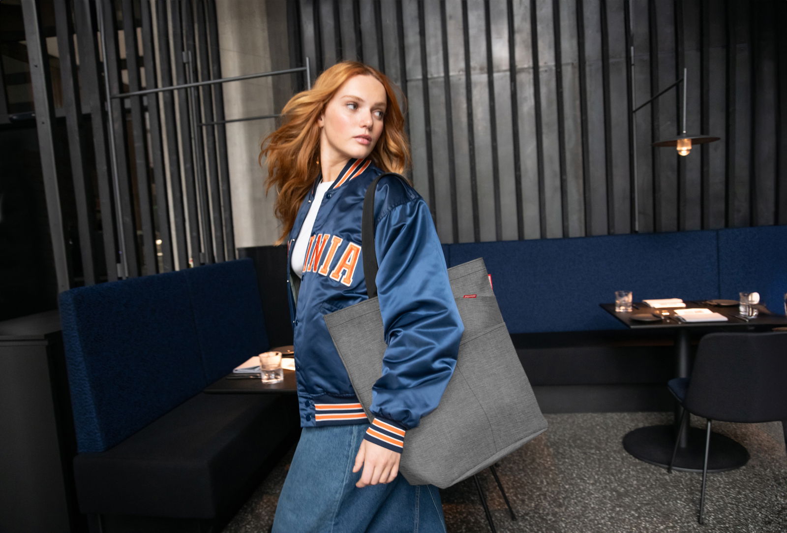 Frau mit blauer Jacke und grauem Cityshopper in einem modernen Café, seitliche Perspektive.