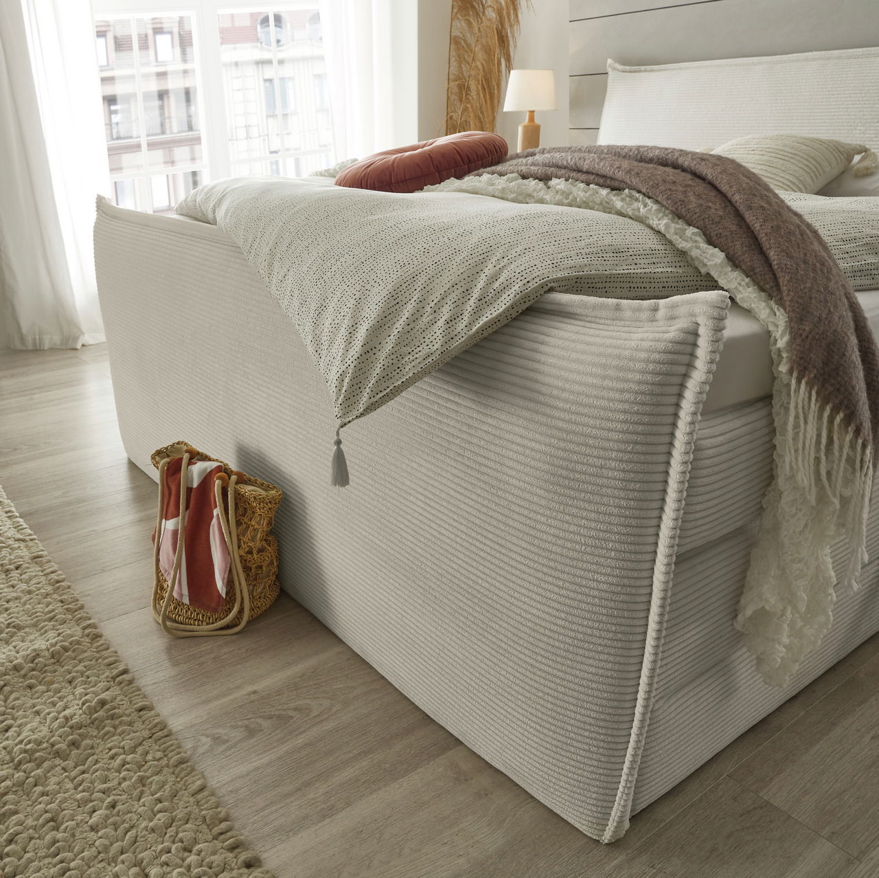 Seitliche Perspektive eines stilvollen Boxspringbetts mit eleganter Bettwäsche und dekorativen Kissen in einem hellen, modernen Schlafzimmer.