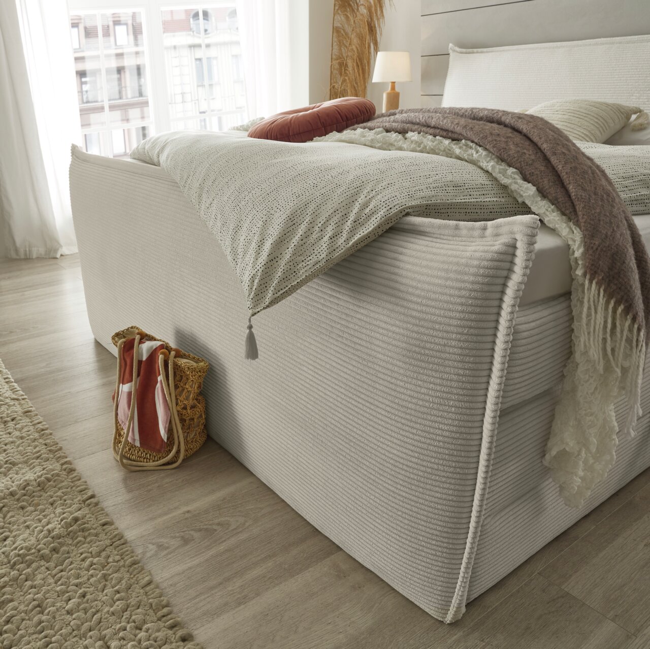 Seitliche Perspektive eines stilvollen Boxspringbetts mit eleganter Bettwäsche und dekorativen Kissen in einem hellen, modernen Schlafzimmer.