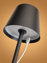 Nahaufnahme einer schwarzen Akku-Tischleuchte von unten mit angeschlossenem Ladekabel, vor einem orangefarbenen Hintergrund.