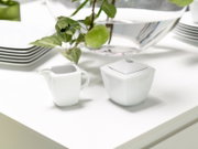 Weißes, eckiges Porzellan-Zucker- und Milchkännchen-Set auf einem hellen Tisch, fotografiert in leichter Seitenansicht von schräg oben; im Hintergrund dekorative Schale mit grünen Äpfeln.