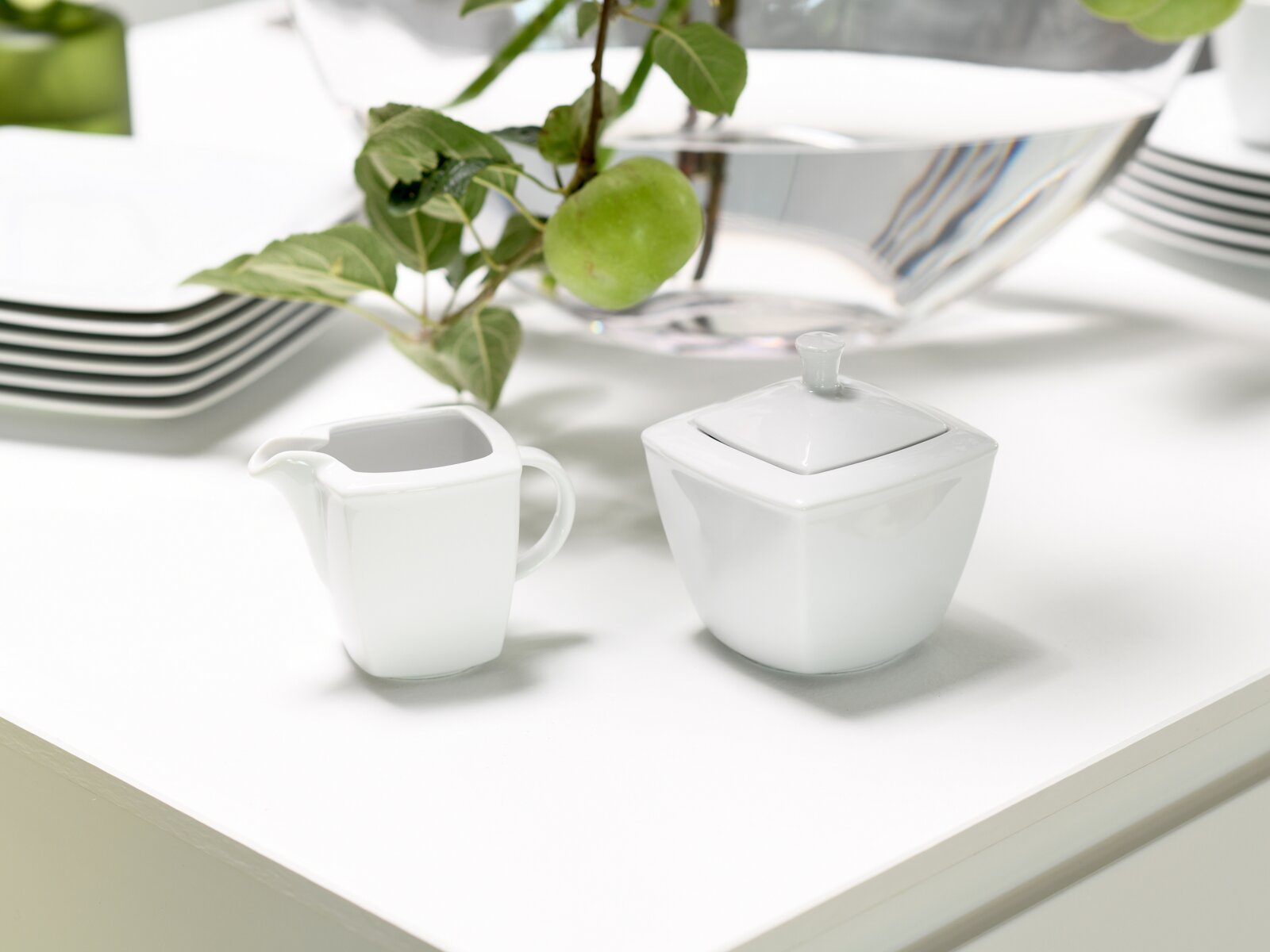 Weißes, eckiges Porzellan-Zucker- und Milchkännchen-Set auf einem hellen Tisch, fotografiert in leichter Seitenansicht von schräg oben; im Hintergrund dekorative Schale mit grünen Äpfeln.