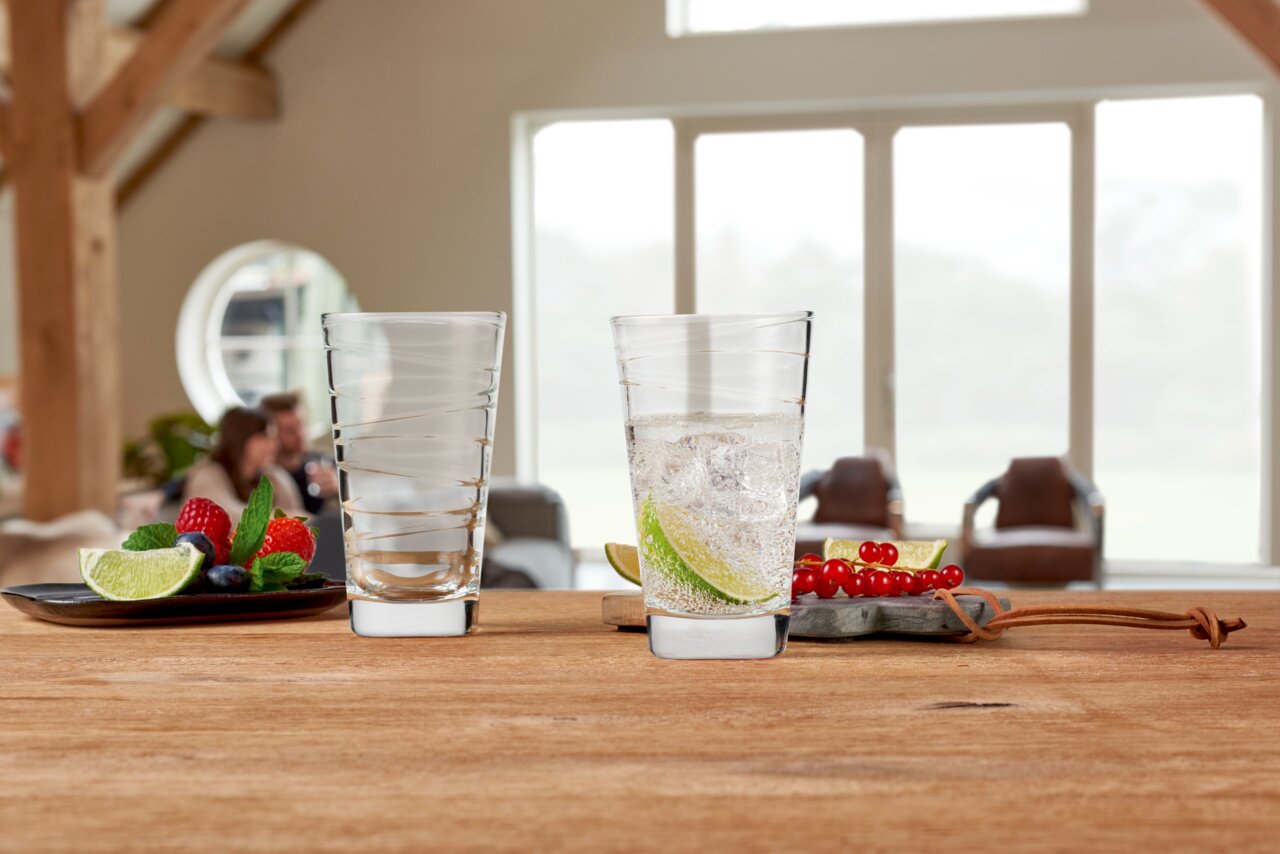 Twee Vario-drinkglazen van 280 ml van Leonardo op een houten tafel, waarvan één gevuld met een drankje en schijfjes limoen, met op de achtergrond een gezellige woonkamer.