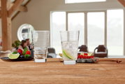 Twee Vario-drinkglazen van 280 ml van Leonardo op een houten tafel, waarvan één gevuld met een drankje en schijfjes limoen, met op de achtergrond een gezellige woonkamer.