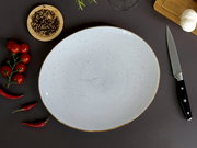 Großer runder, hellgrau gesprenkelter Keramikteller mit braunem Rand auf dunkler Arbeitsfläche, fotografiert in Draufsicht; daneben Messer, Knoblauch, Chilis, Kirschtomaten und Schale mit Pfefferkörnern.