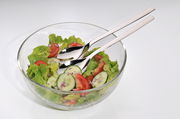 Glas-Salatschüssel mit frischem Salat, Gurken, Tomaten und Zwiebeln, serviert mit glänzendem Salatbesteck. Ansicht von oben.
