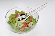 Glas-Salatschüssel mit frischem Salat, Gurken, Tomaten und Zwiebeln, serviert mit glänzendem Salatbesteck. Ansicht von oben.