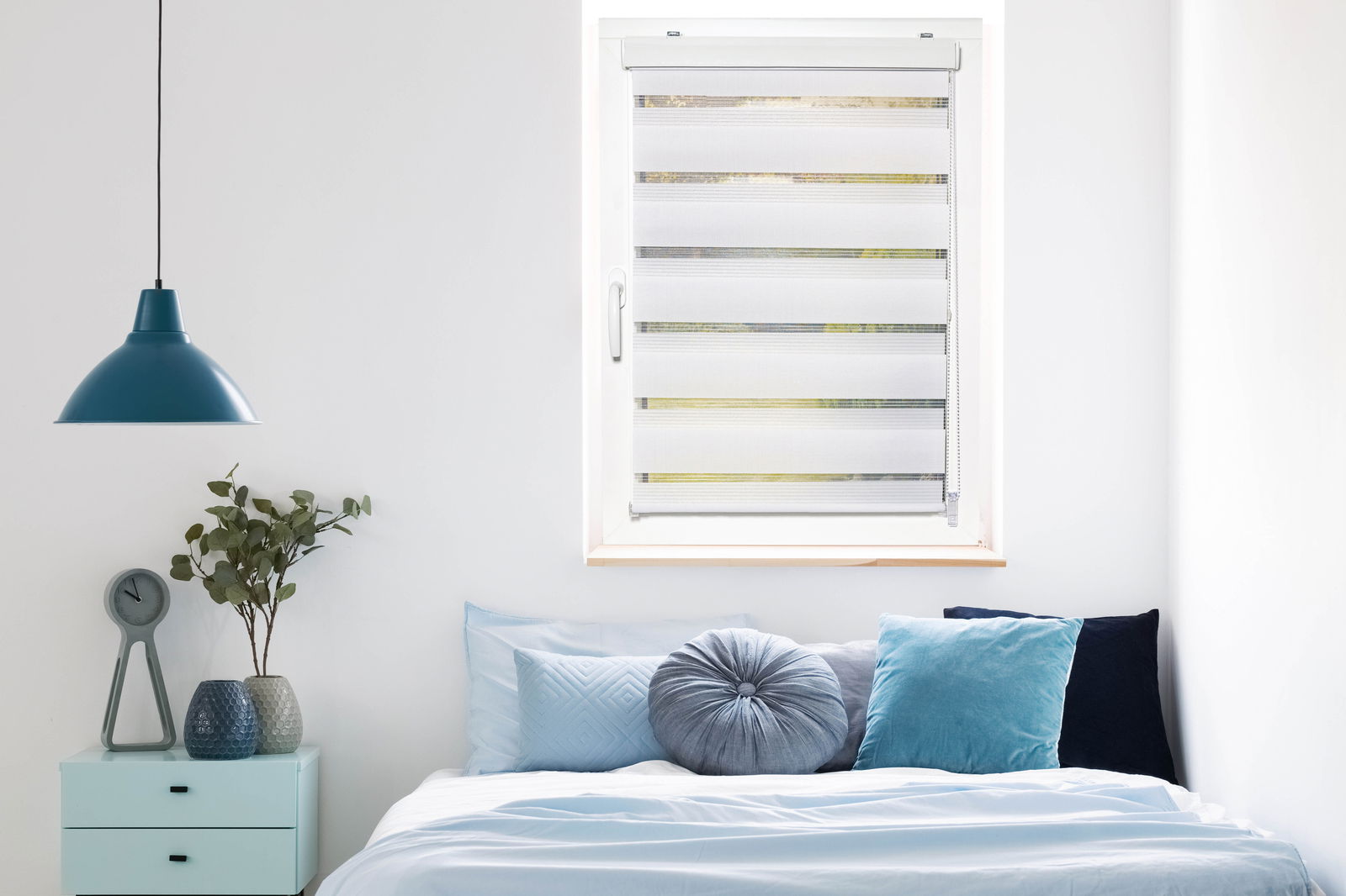 Doppelrollo mit Blende in einem modernen Schlafzimmer, frontal betrachtet, mit einem Bett und dekorativen Kissen darunter.
