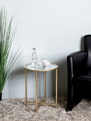 Runder Beistelltisch mit goldfarbenem Metallgestell und Glasplatte neben schwarzem Ledersessel, frontale Perspektive im Wohnraum