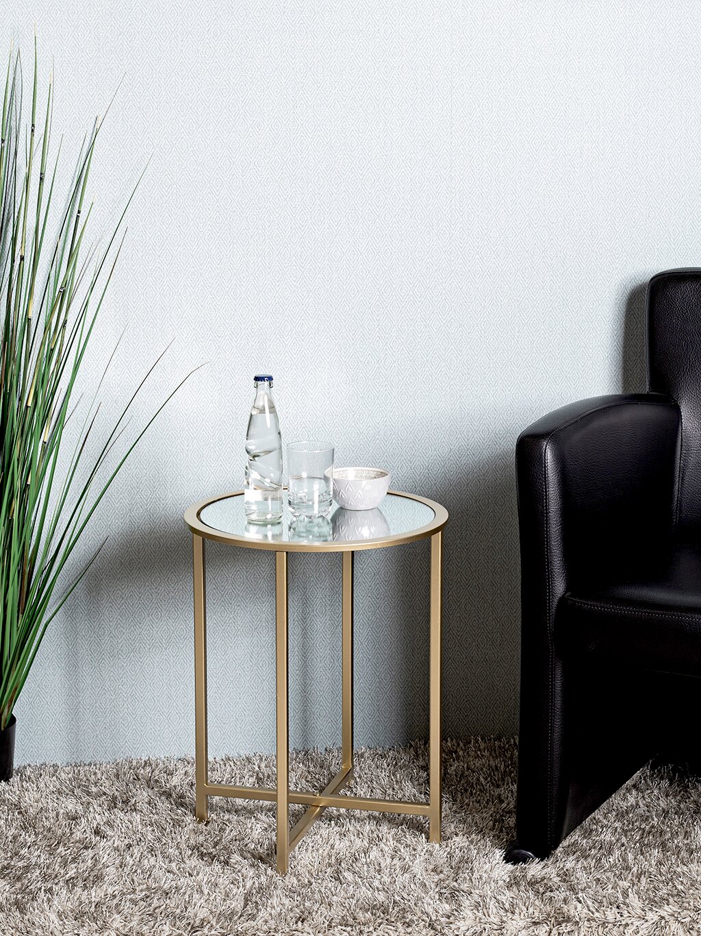 Runder Beistelltisch mit goldfarbenem Metallgestell und Glasplatte neben schwarzem Ledersessel, frontale Perspektive im Wohnraum