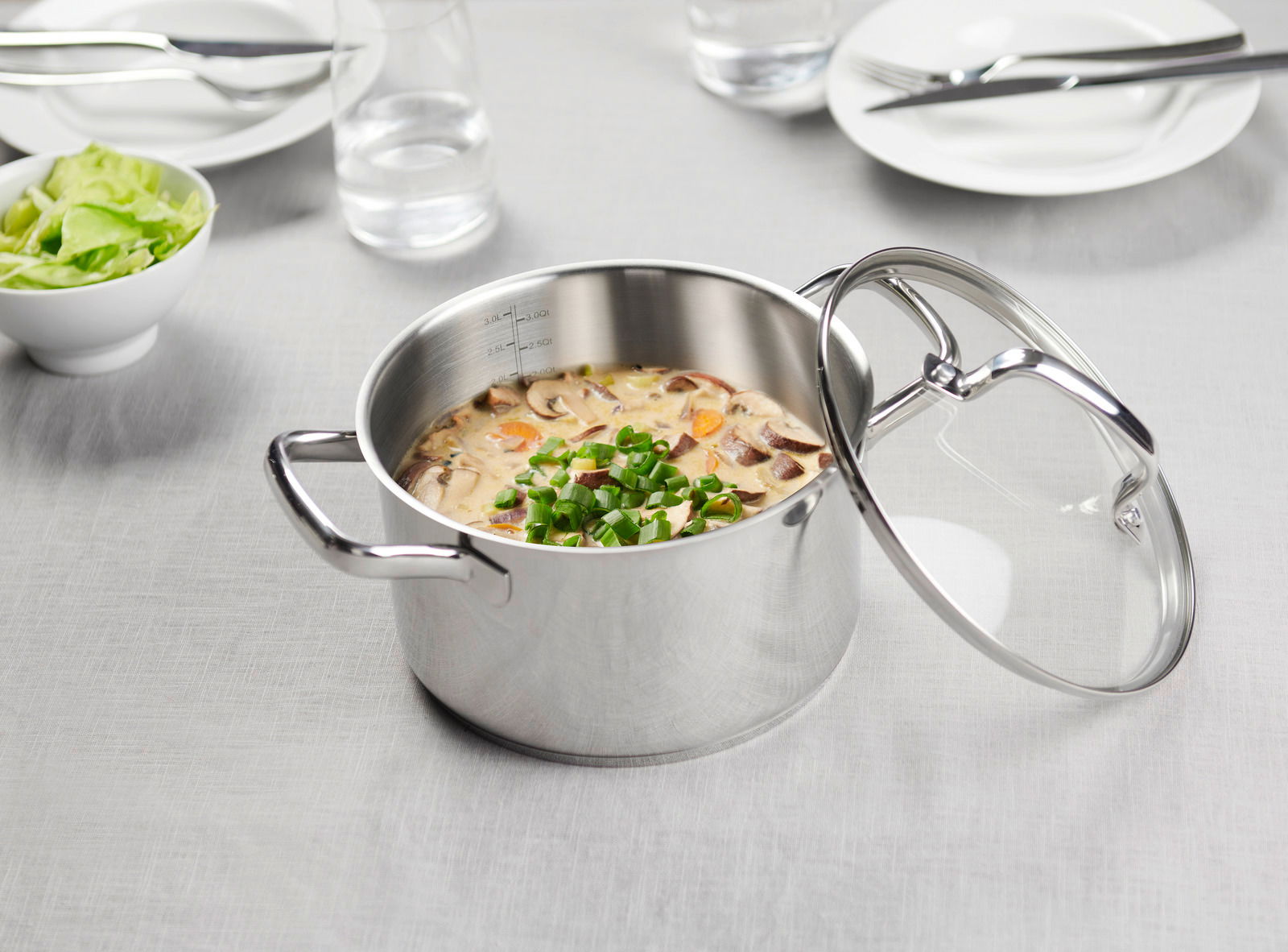 Kochtopf aus Edelstahl mit Glasdeckel, gefüllt mit einer Suppe, auf einem gedeckten Tisch aus der Vogelperspektive.