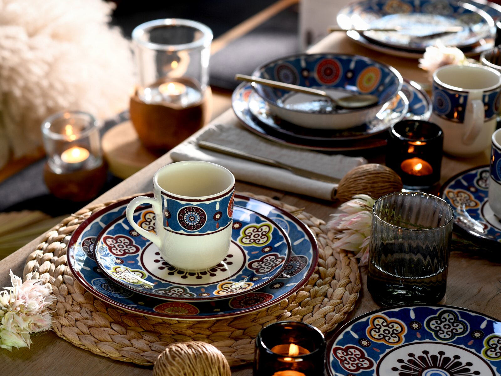 Schräg von oben fotografiertes Tischgedeck mit blau gemustertem Keramikgeschirr: Tasse auf Teller, Schale mit Löffel, Kerzen und Glas auf Holztisch mit geflochtenem Platzset.