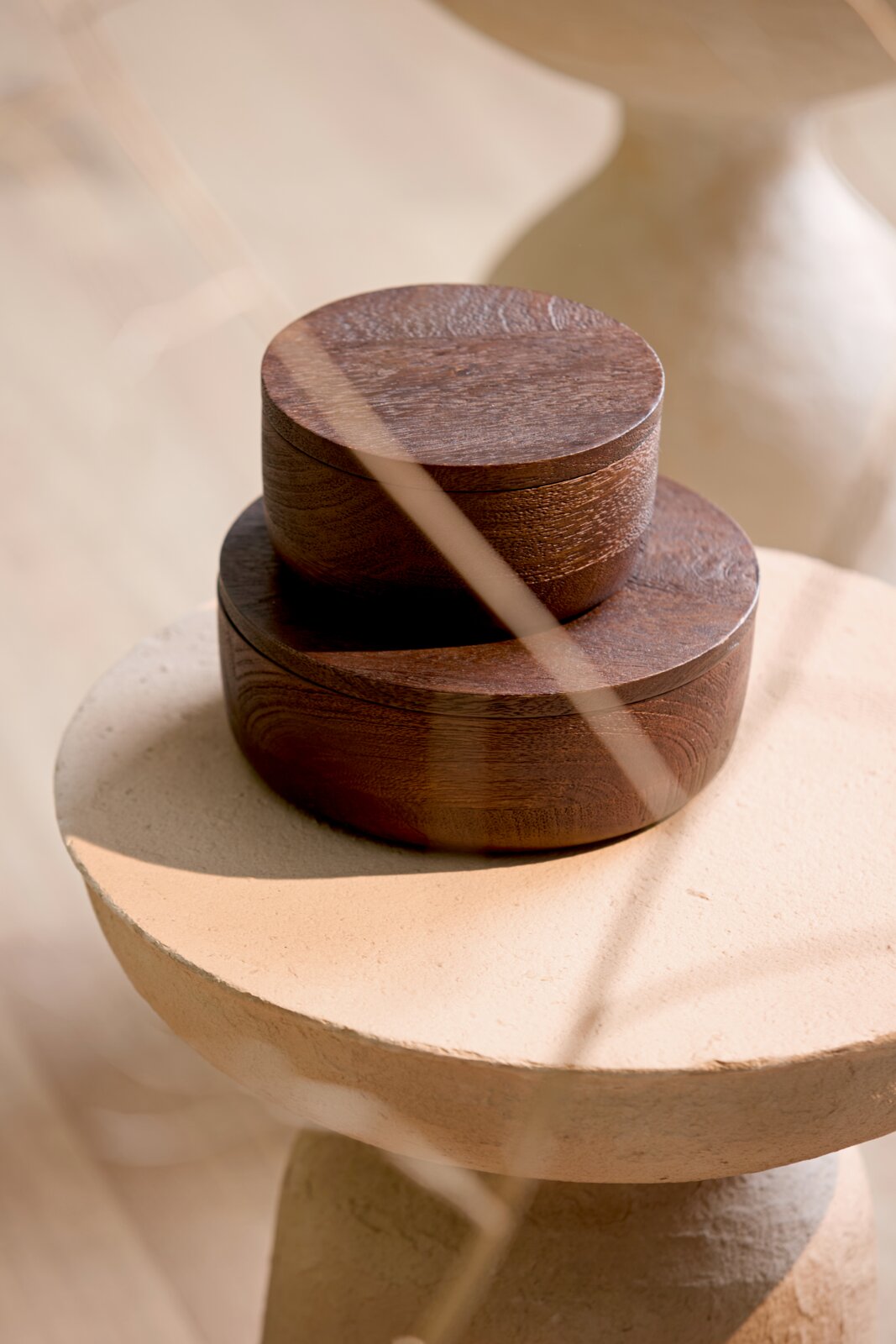 Zwei runde Dosen aus dunkelbraunem Holz mit Deckel, übereinander arrangiert auf einem hellen Beistelltisch, aus schräger Vorderansicht fotografiert.