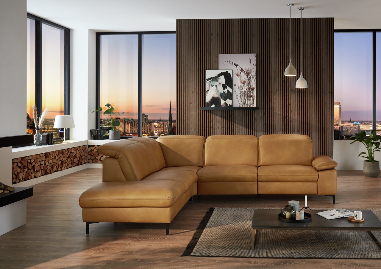 Beldomo Premium Ecksofa PAMPLONA Moderne Polsterrundecke in einem stilvollen Wohnzimmer mit Blick auf die Stadt bei Sonnenuntergang, aus einer frontalen Perspektive aufgenommen.