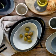 Draufsicht auf modern gedeckten Tisch mit ovalen Keramikschalen und Tellern in Blau- und Beigetönen, Oliven in einer Schale sowie Öl, Salz und Brot auf Holzuntergrund