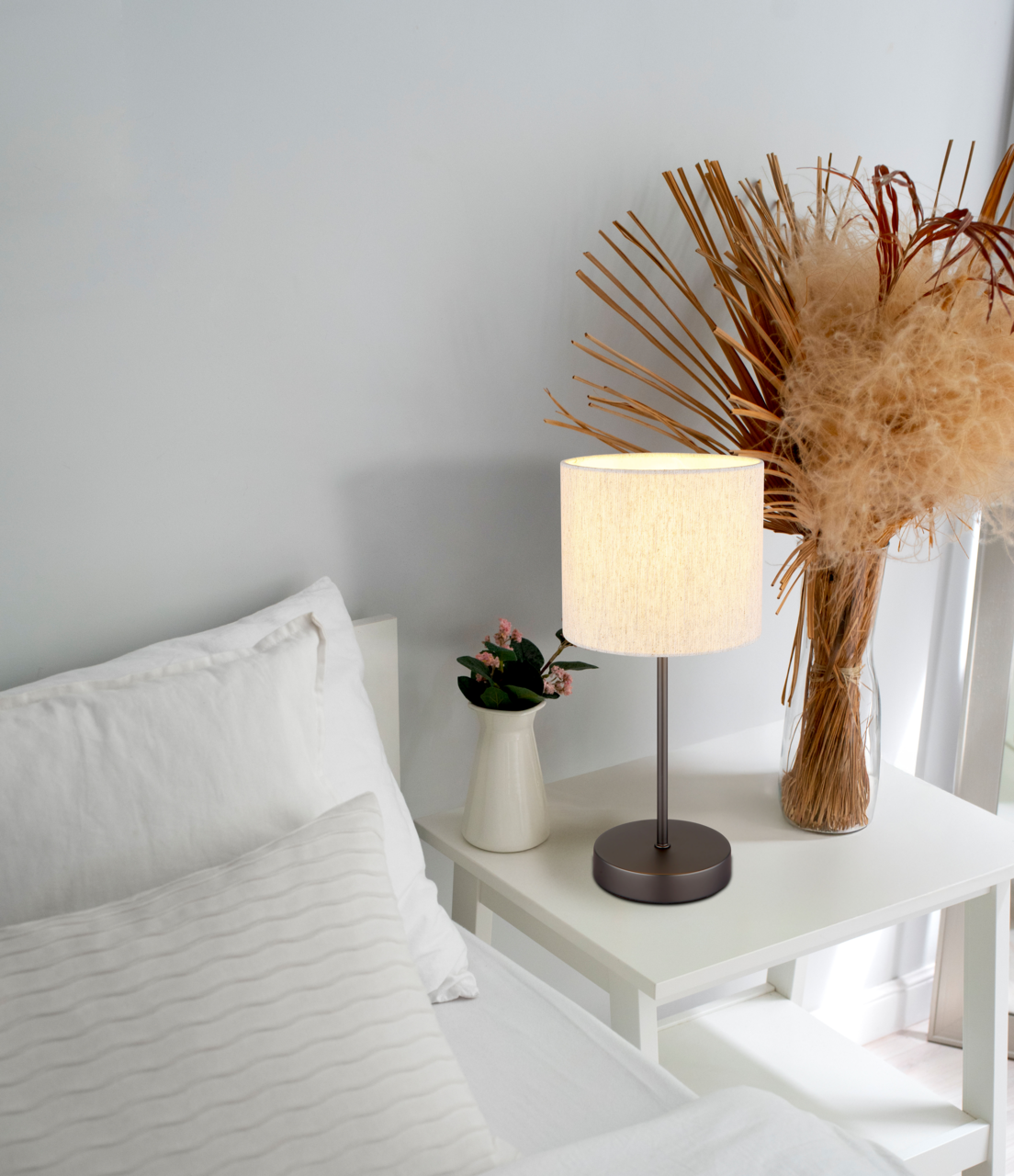 Tischleuchte auf einem Nachttisch neben einem Bett, mit einem weißen Lampenschirm und einem dunklen Sockel, umgeben von Dekorationen wie einer Vase mit Blumen und getrockneten Pflanzen. Perspektive von der Seite.
