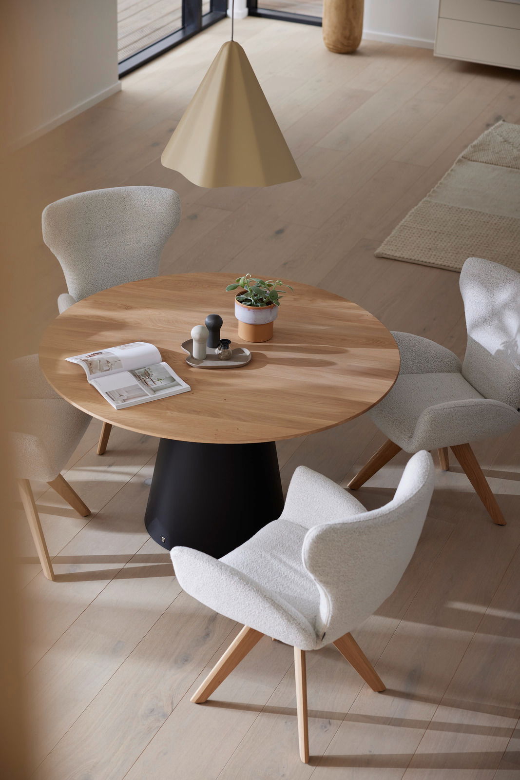 Runder Esstisch mit Holzplatte und schwarzem Sockel, umgeben von vier gepolsterten Armlehnstühlen in einem modernen Esszimmer, aus der Vogelperspektive.