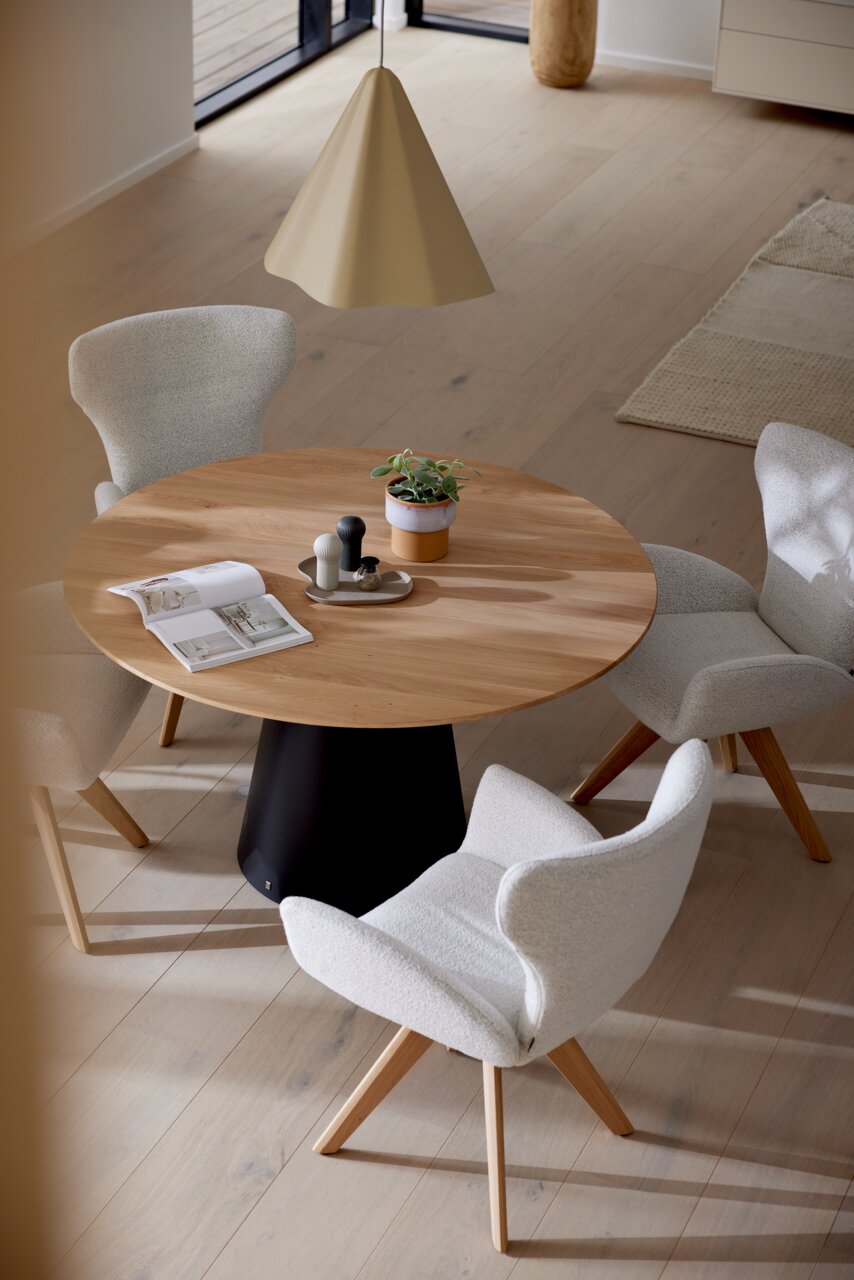 Runder Esstisch mit Holzplatte und schwarzem Sockel, umgeben von vier gepolsterten Armlehnstühlen in einem modernen Esszimmer, aus der Vogelperspektive.