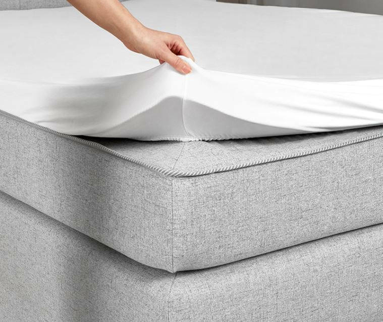 Nahaufnahme eines grauen Boxspringbetts mit einer Hand, die ein weißes Spannbettlaken aufzieht.