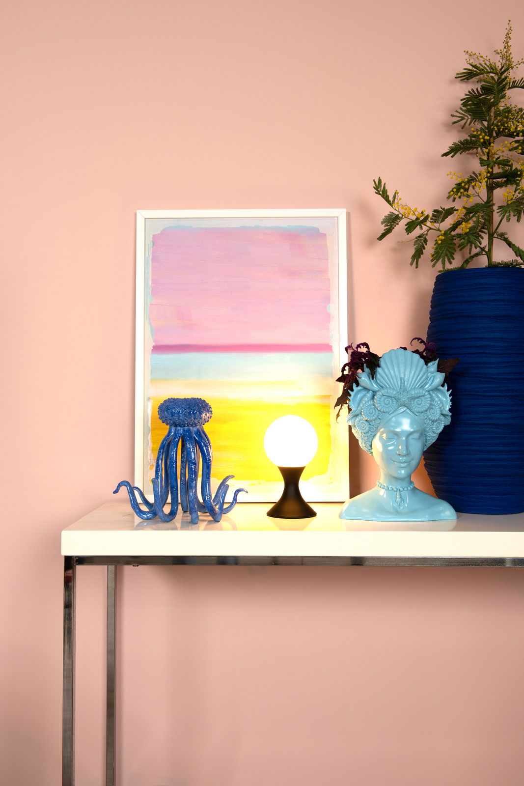 Moderne Tischleuchte MOYA mit kugelförmigem Leuchtkörper auf einem Tisch, umgeben von dekorativen Objekten, seitliche Perspektive.