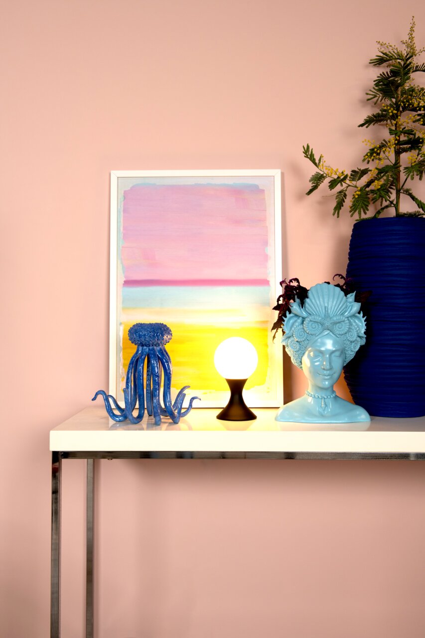 Lucide Tischleuchte MOYA Moderne Tischleuchte MOYA mit kugelförmigem Leuchtkörper auf einem Tisch, umgeben von dekorativen Objekten, seitliche Perspektive.