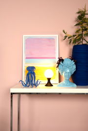 Moderne Tischleuchte MOYA mit kugelförmigem Leuchtkörper auf einem Tisch, umgeben von dekorativen Objekten, seitliche Perspektive.