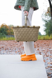 Seitenansicht eines Carrybag Rhombus Olive, getragen von einer Person in weißer Hose und orangefarbenen Schuhen, im Freien auf einem herbstlichen Weg.