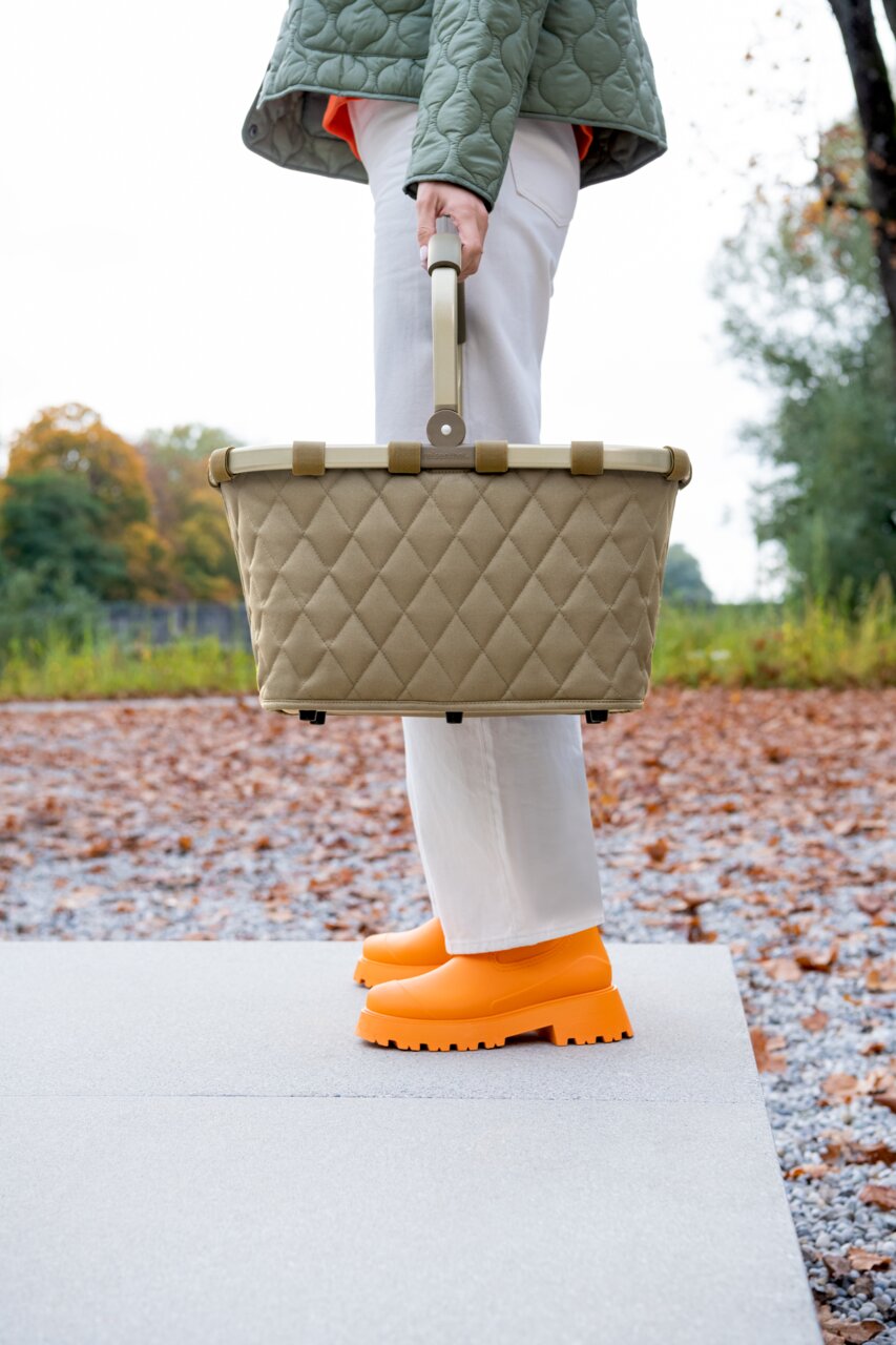 Zijaanzicht van een Carrybag Rhombus Olive, gedragen door iemand in een witte broek en oranje schoenen, buiten op een herfstpad.