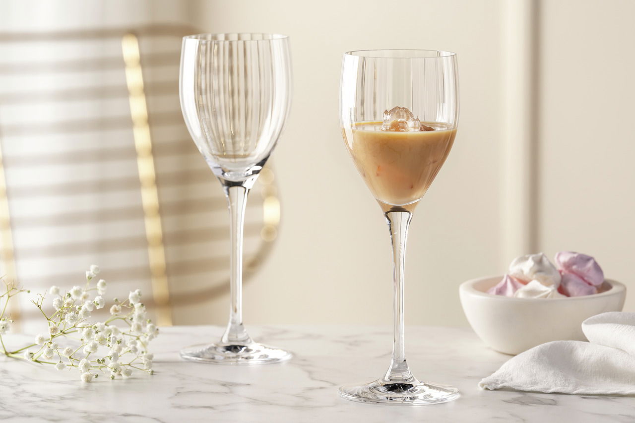 Leonardo Likörglas POESIA Zwei elegante Likörgläser POESIA mit feinen vertikalen Rillen, eines gefüllt mit einem cremigen Getränk und Eiswürfeln, auf einem Marmortisch mit zarten weißen Blüten und einer Schale mit bunten Süßigkeiten im Hintergrund. Seitliche Perspektive.