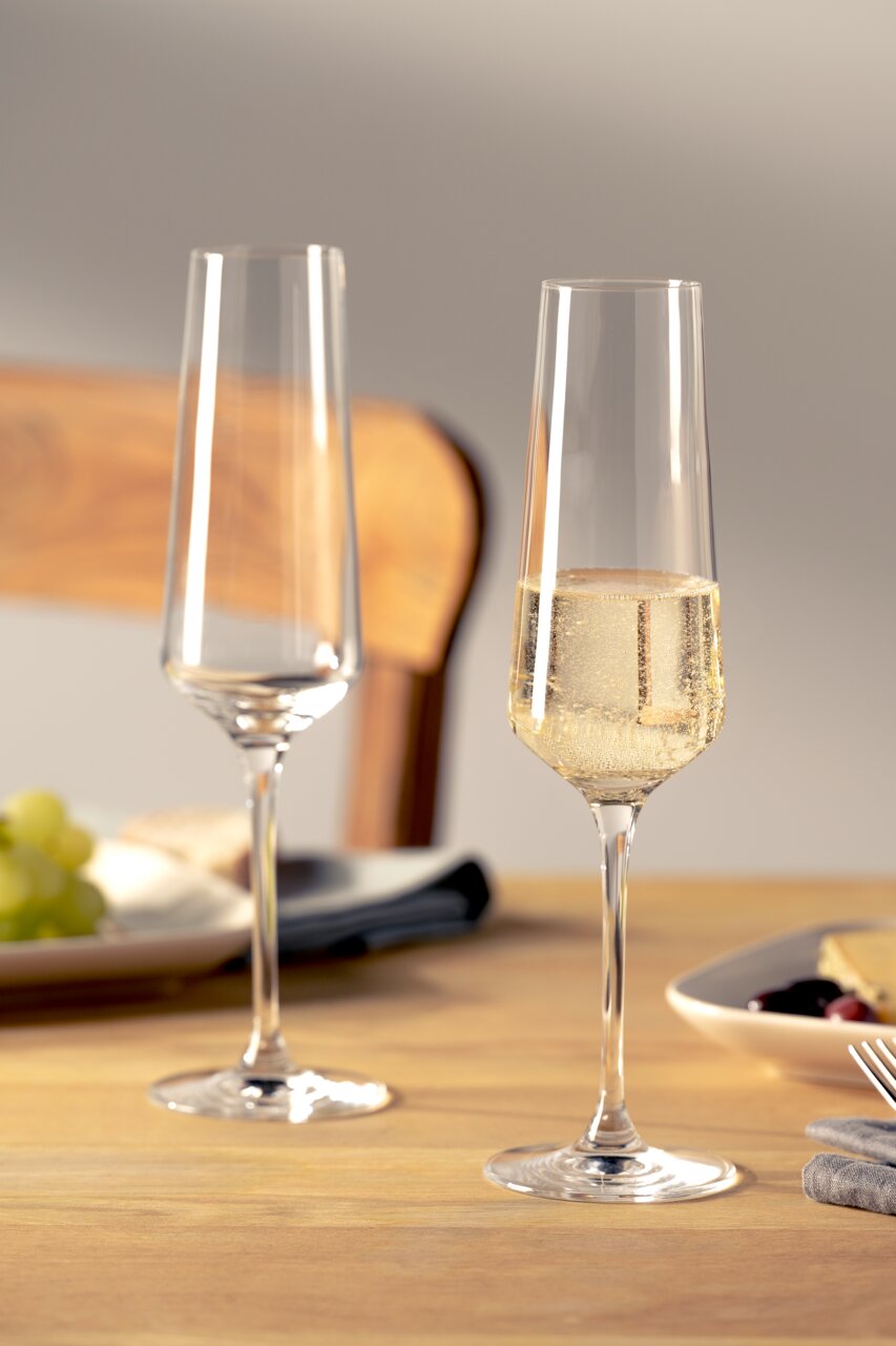Zwei elegante Sektgläser auf einem gedeckten Tisch, eines gefüllt mit sprudelndem Sekt, aus seitlicher Perspektive fotografiert.