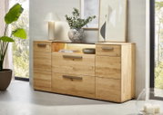 Moderne Holz-Kommode aus hellem Massivholz mit Metallgriffen und offener Ablage, dekoriert mit Lampe, Vase und Bildern, frontal in leicht schräger Perspektive von vorn fotografiert.
