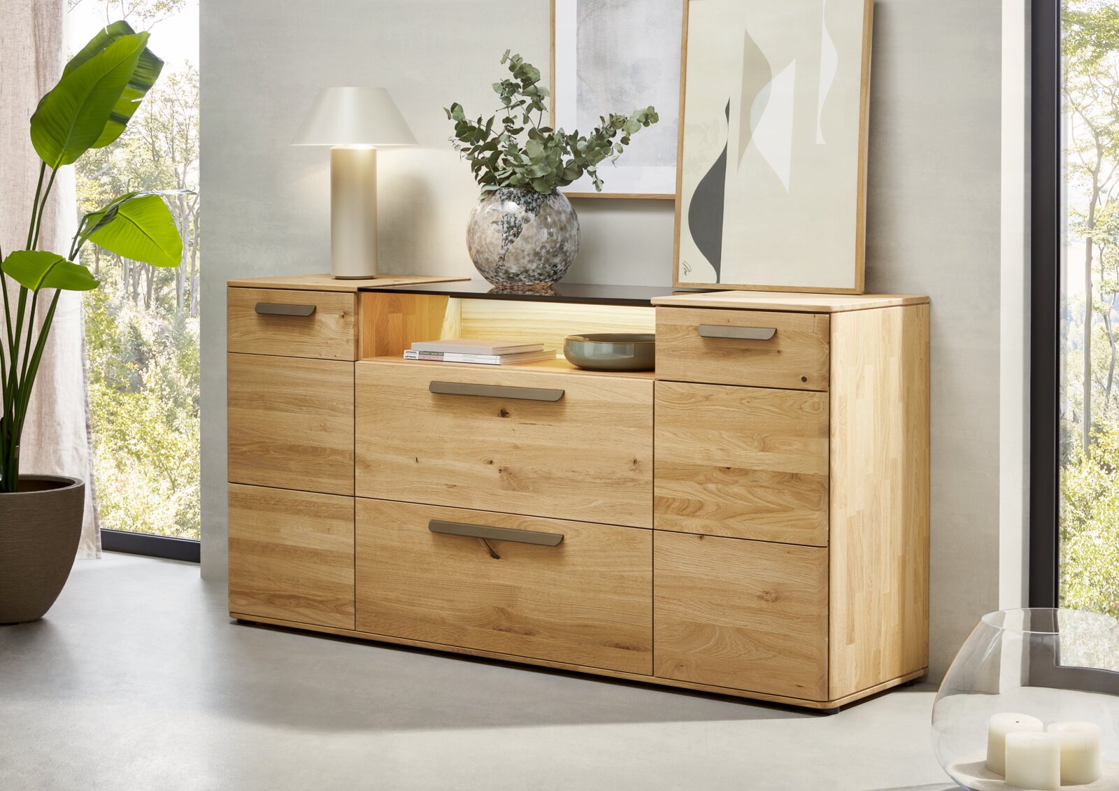 Moderne Holz-Kommode aus hellem Massivholz mit Metallgriffen und offener Ablage, dekoriert mit Lampe, Vase und Bildern, frontal in leicht schräger Perspektive von vorn fotografiert.