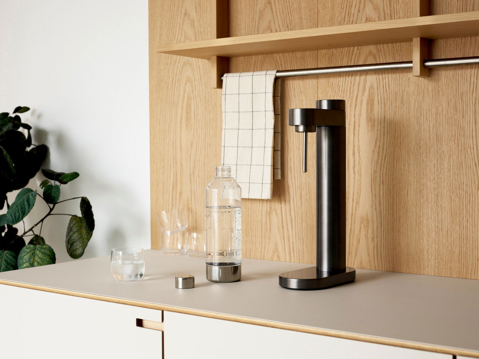 Schwarzer Wassersprudler auf einer Küchenarbeitsplatte, seitliche Perspektive, mit einer gefüllten Flasche und einem Glas Wasser daneben.