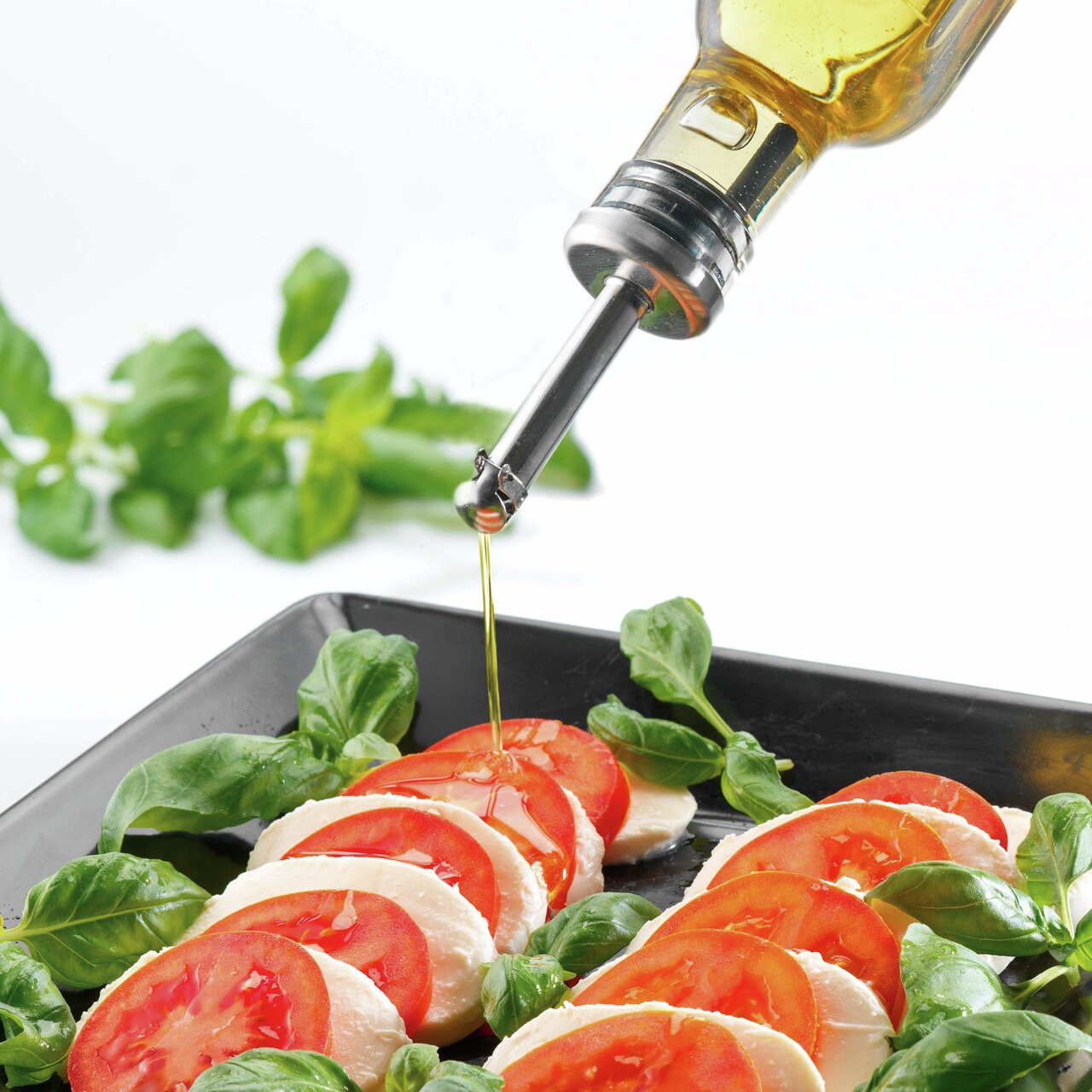 Nahaufnahme eines Ausgießers, der Öl auf einen frischen Tomaten-Mozzarella-Salat mit Basilikumblättern träufelt.