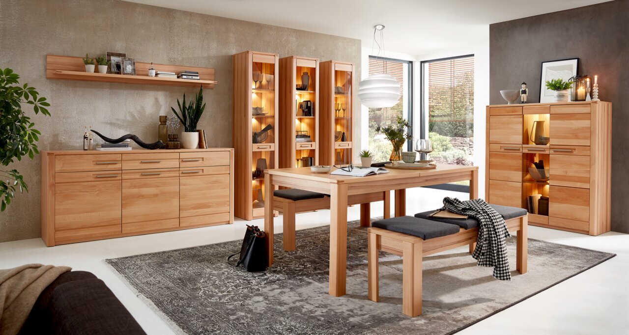 Wohnzimmer mit modernen Holzmöbeln, darunter ein Sideboard, Vitrinen und ein Esstisch mit Bänken, aus der Perspektive eines offenen Raumes mit viel Tageslicht.