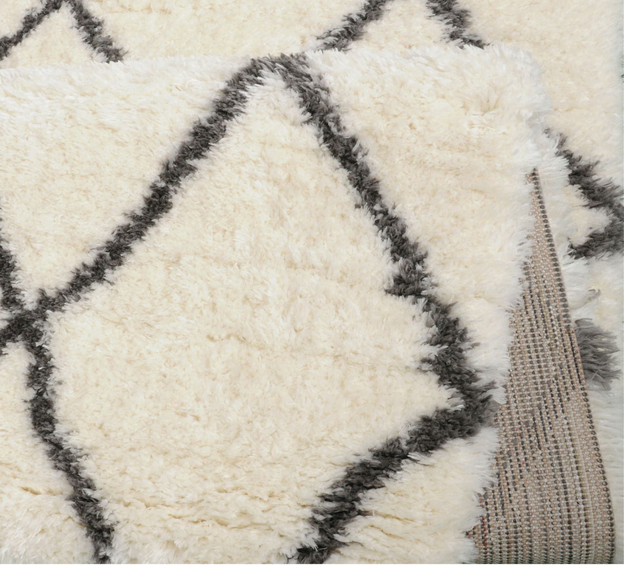 Nahaufnahme eines flauschigen, cremefarbenen Teppichs mit grauem Rautenmuster, teilweise aufgerollt.