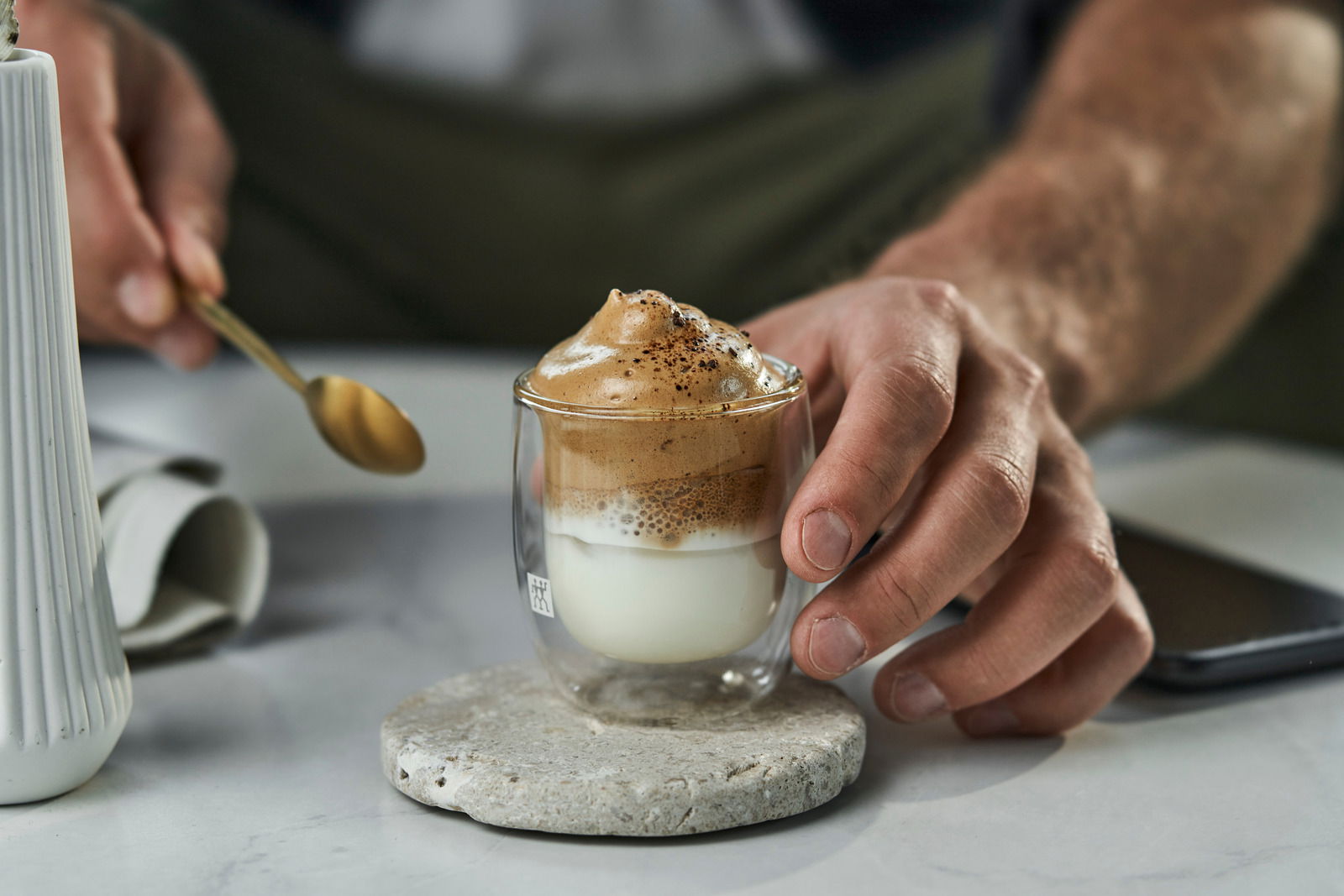 Nahaufnahme eines Espressoglases mit doppelwandigem Design, gefüllt mit einem Schichtgetränk aus Milch und Kaffee, auf einem runden Steinuntersetzer. Eine Hand hält das Glas, während eine andere Hand einen goldenen Löffel hält.