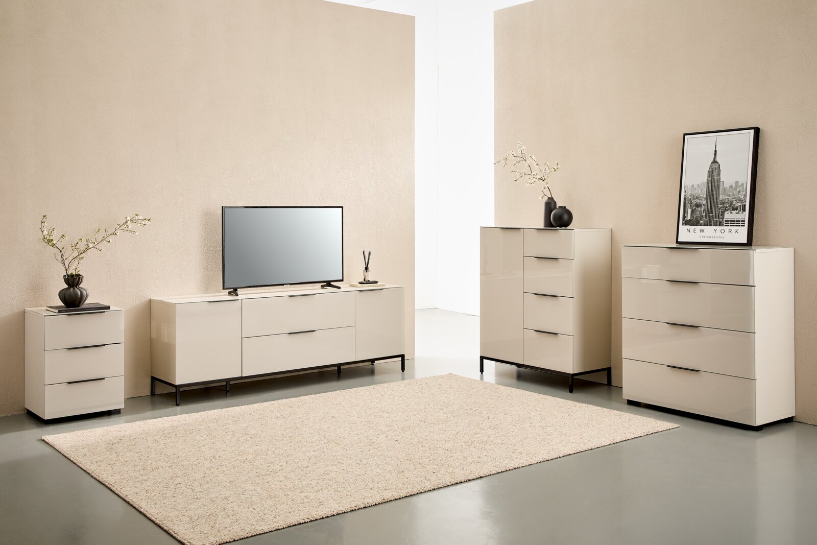 Modernes Wohnzimmer-Set mit cremefarbenen Hochglanz-Kommoden und TV-Lowboard auf schwarzen Metallfüßen, dekoriert mit Vase, Bild und Fernseher, gezeigt in frontaler Perspektive mit leichtem Weitwinkel.
