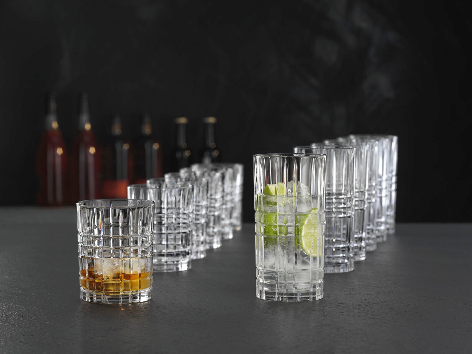 Ein Set von zwölf Kristallgläsern der Serie Highland, auf einer dunklen Oberfläche arrangiert. Im Vordergrund ein kurzes Glas mit Whisky und ein hohes Glas mit Gin Tonic und Limettenscheiben. Perspektive von vorne.