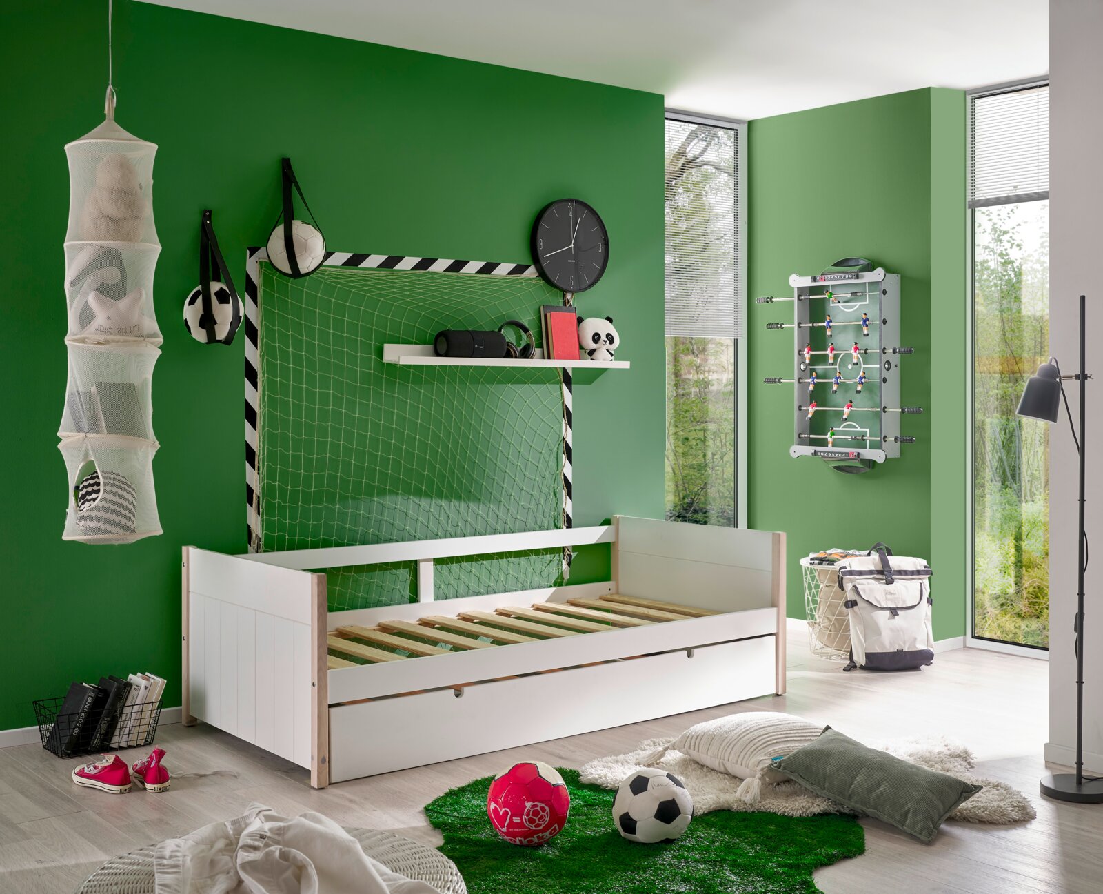 Weißes Jugendbett in einem sportlich dekorierten Zimmer mit grüner Wand, Fußball-Dekorationen und einem Tischkicker an der Wand. Perspektive: seitlich von vorne.
