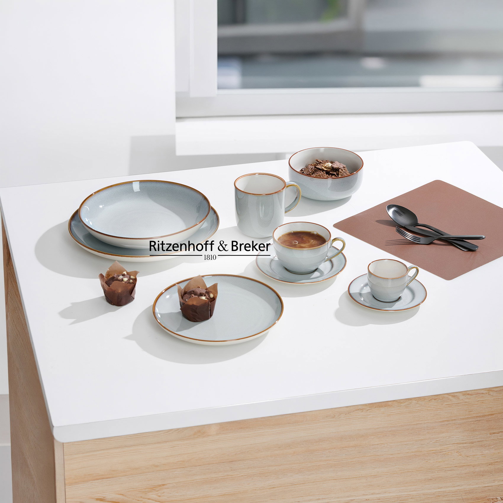 Grijs keramisch servies met kopjes, kommen en borden op witte tafel, met koffie en muffins.
