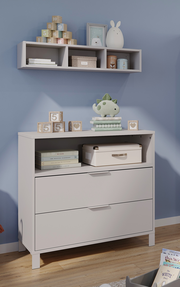 Weiße, schmale Kommode mit zwei Schubladen und einem offenen Fach, dekoriert mit Spielzeug und Büchern, in einem Kinderzimmer. Perspektive von vorne.