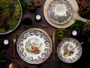 Festlich gedeckter Tisch mit braun-weiß gemustertem Porzellangeschirr (Teller mit Hirsch- und Fuchsmotiv), Tasse, grünen Gläsern und goldfarbenem Besteck, dekoriert mit Tannenzweigen, Zapfen und Kerzen; Perspektive: Draufsicht von oben (Flatlay).