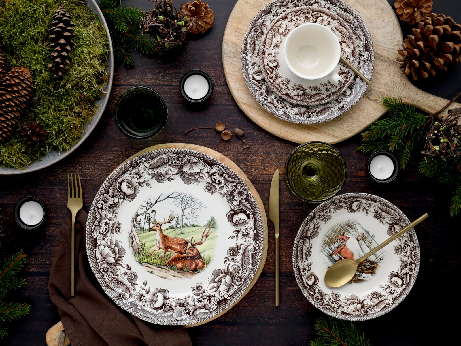 Festlich gedeckter Tisch mit braun-weiß gemustertem Porzellangeschirr (Teller mit Hirsch- und Fuchsmotiv), Tasse, grünen Gläsern und goldfarbenem Besteck, dekoriert mit Tannenzweigen, Zapfen und Kerzen; Perspektive: Draufsicht von oben (Flatlay).