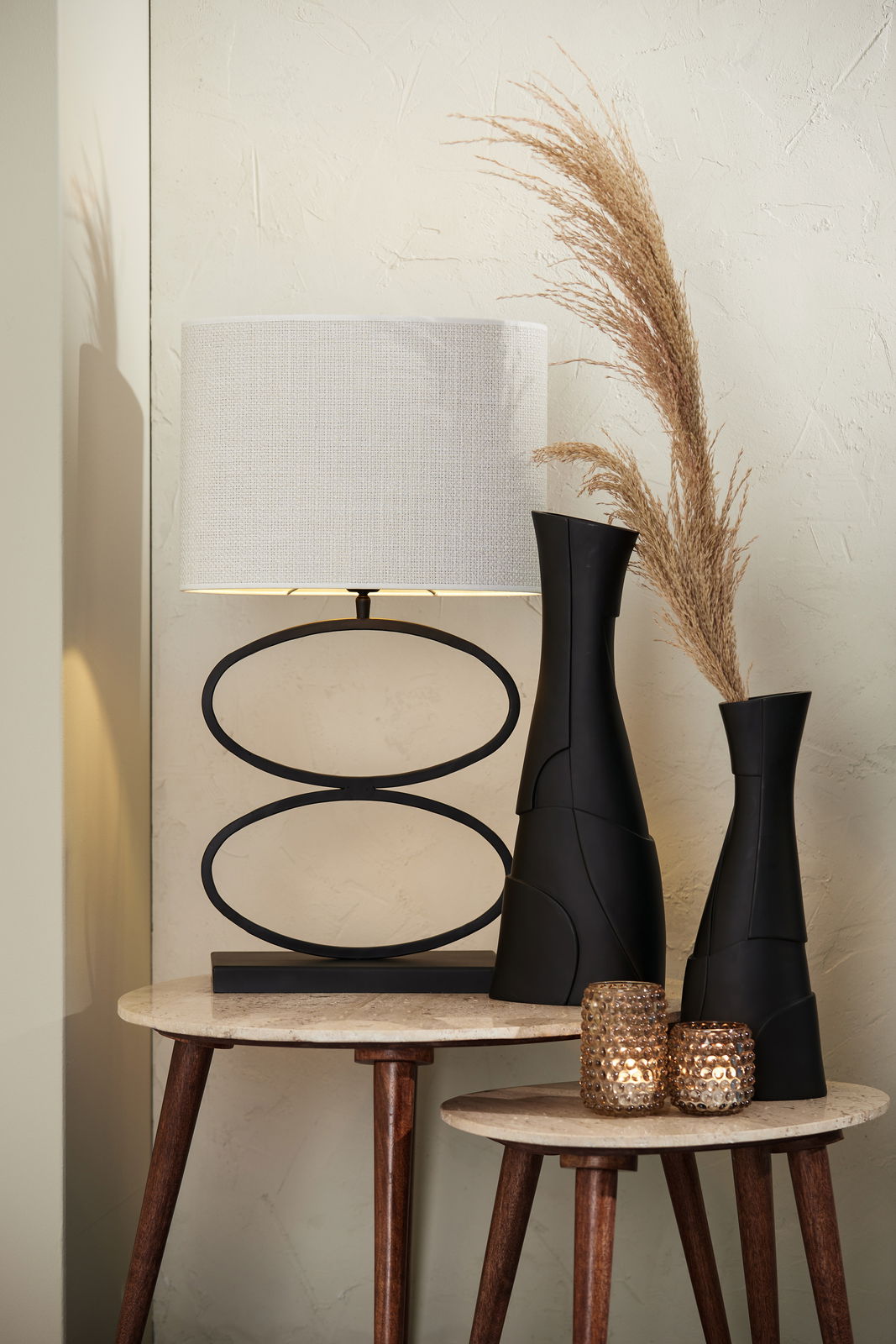 Moderner ovaler Lampenschirm in hellem Beige auf einem stilvollen schwarzen Metallfuß, präsentiert auf einem Tisch mit dekorativen Vasen und Kerzenhaltern, seitliche Perspektive.