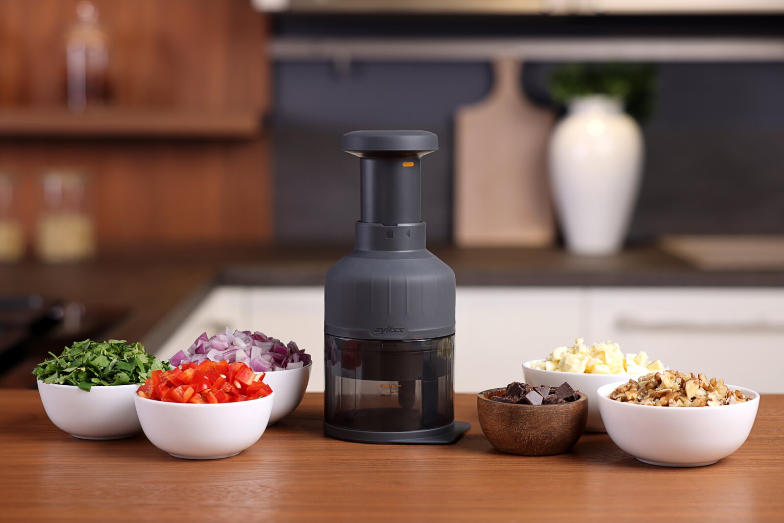 Zyliss Groentehakker ZICK ZACK 3 Vooraanzicht van een zwarte handmatige hakmolen op een houten aanrecht, omringd door schaaltjes met gehakte kruiden, paprika, rode ui, kaas, chocolade en noten in een keukenomgeving.